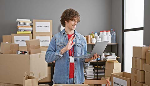 Department of Veterans Affairs Voluntary Service (VAVS) National Advisory Committee - A friendly young man with curly hair stands amid cardboard boxes in a warehouse, using a laptop and gesturing.