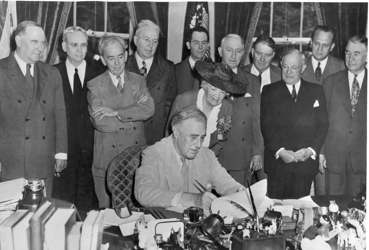 Edith Rogers leans in as President Franklin Roosevelt signs the GI Bill on June 22, 1944. Moments later, Roosevelt handed her the signing pen as a token of appreciation for her co-sponsorship of the bill. President Roosevelt is sitting in the Oval Office, Rogers directly behind him standing, with a group of 10 men behind her. (FDR Library)